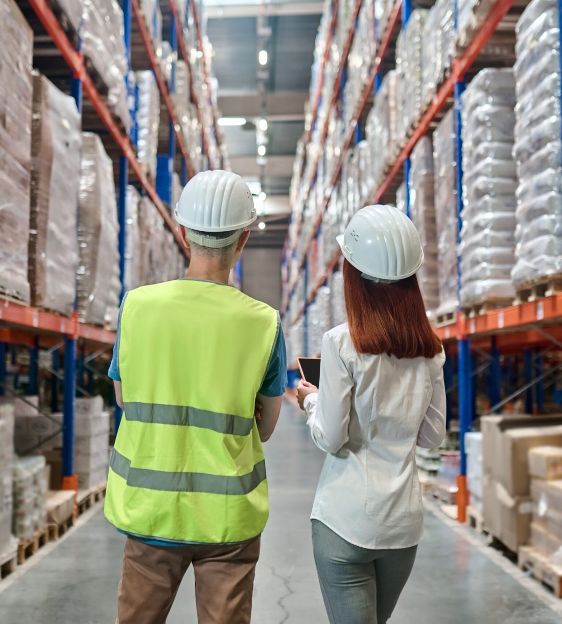 Work moment. Back view of elegant long-haired redhead woman and man in helmet and overalls standing communicating in aisle between warehouse shelves